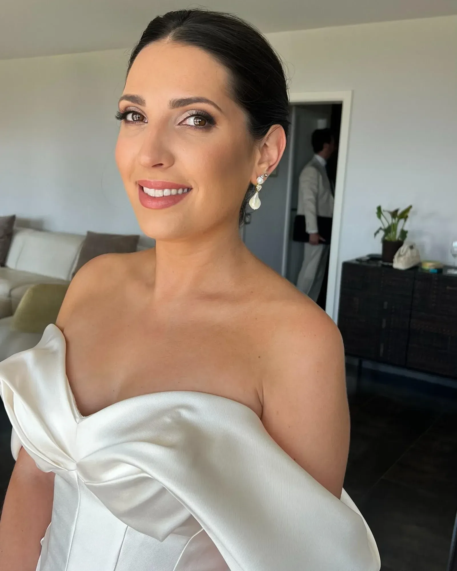 Une femme en tenue de soirée, portant des boucles d'oreilles, souriante, debout dans une pièce où se trouvent un canapé, des coussins, une plante en pot et une personne à l'arrière-plan.