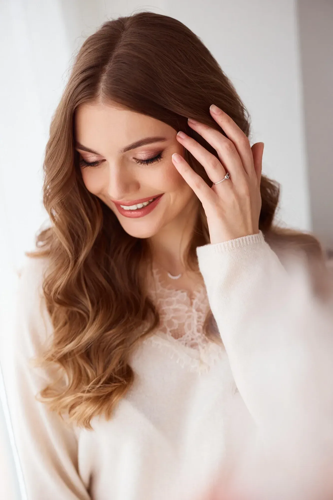 Gros plan sur une femme aux longs cheveux ondulés d'un châtain roux, qui sourit les yeux fermés tout en caressant ses cheveux de la main ; elle porte un pull blanc orné de dentelle.