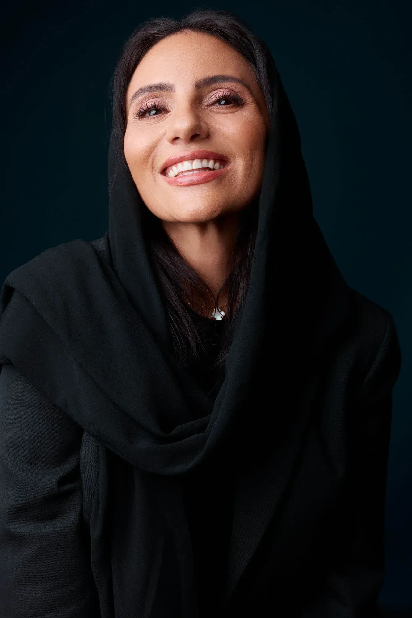 Une femme souriante aux cheveux noirs portant un foulard noir et des vêtements noirs sur un fond sombre.