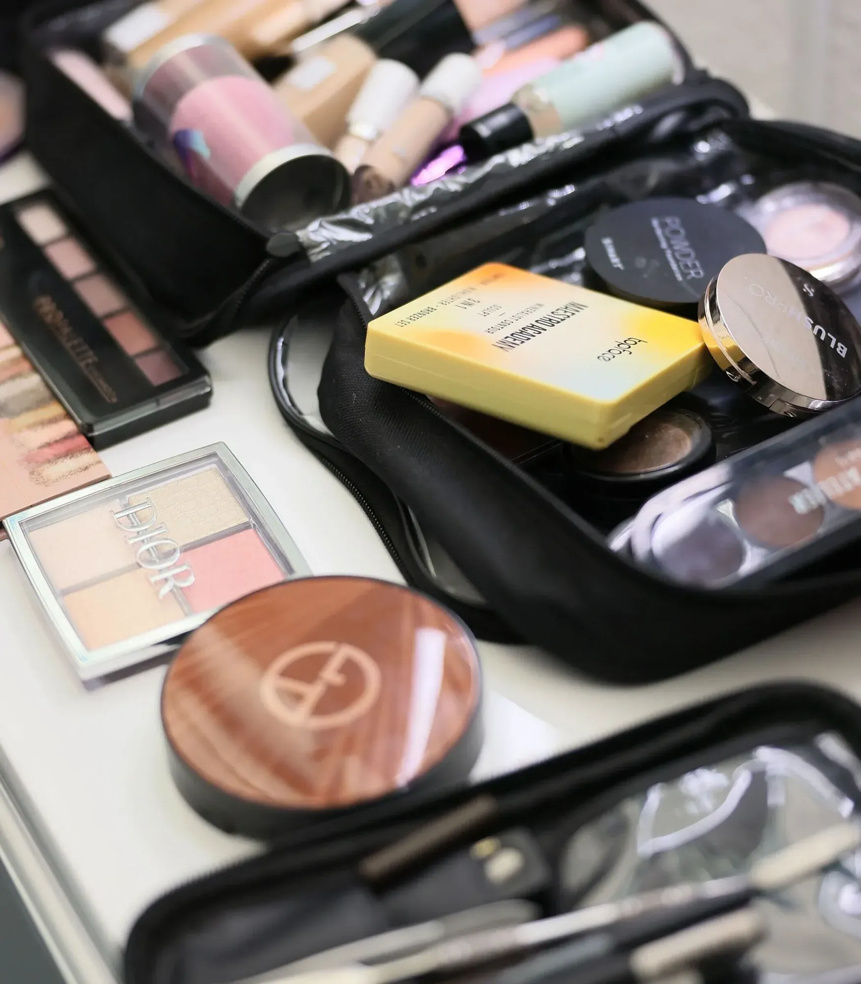 Trousse de maquillage ouverte contenant divers produits cosmétiques, notamment des flacons de fard à joues, de spray fixant et de fond de teint, sur une surface blanche.