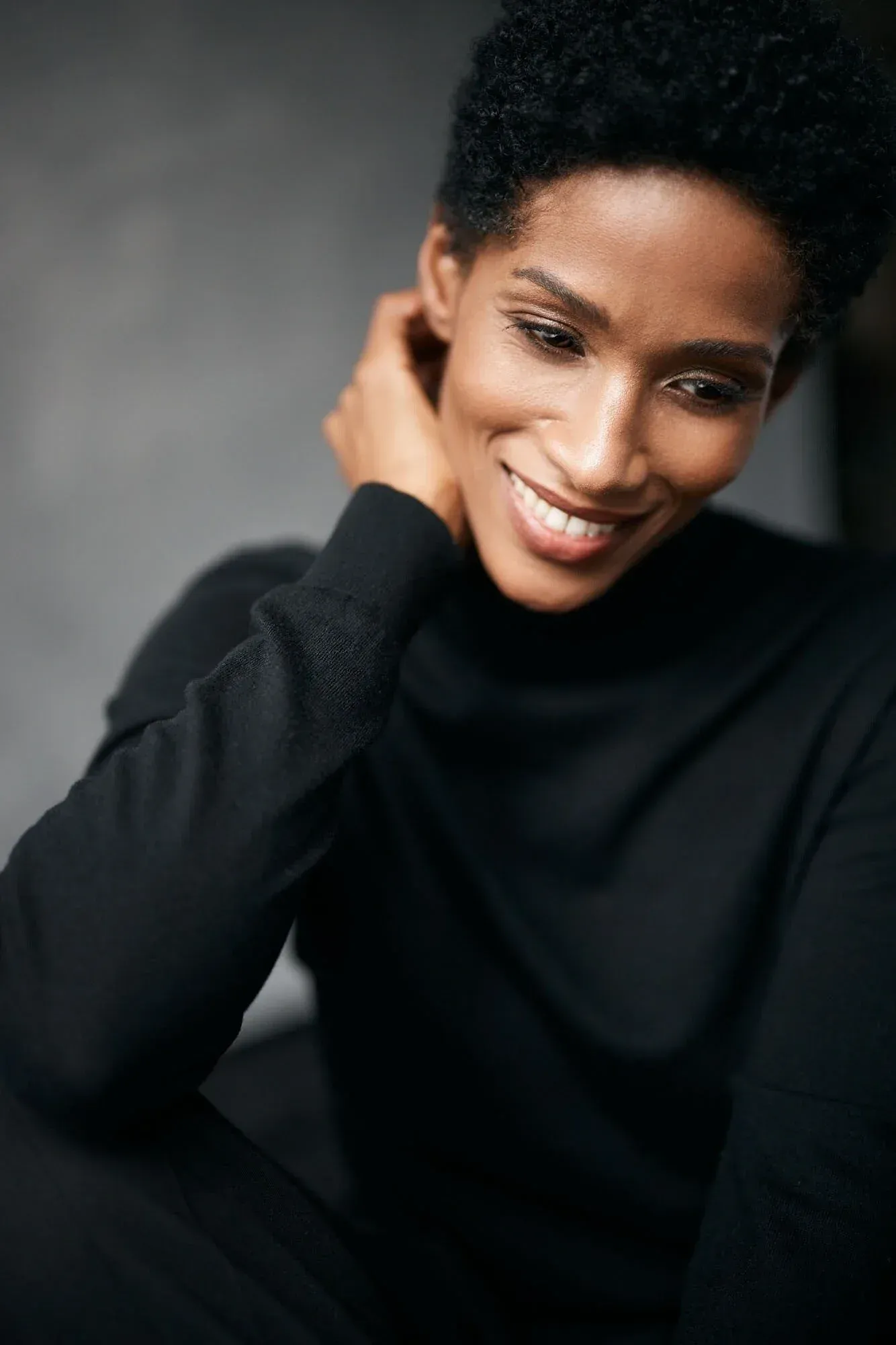 Une jeune femme souriante, vêtue d'un pull à col roulé noir, aux cheveux courts et bouclés, qui se touche la nuque et baisse les yeux, sur fond gris.