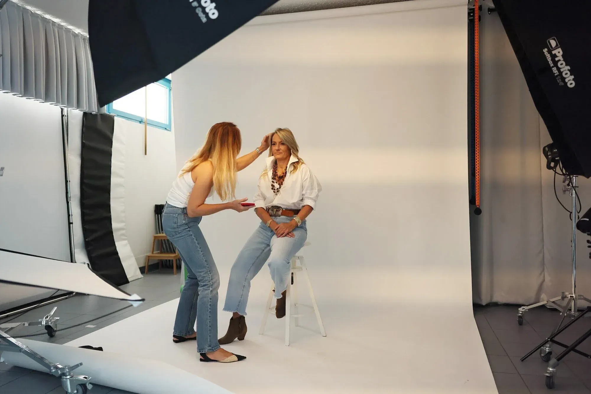 Séance de photos dans un studio : une femme aux cheveux blonds bicolores, avec une chemise blanche et un jean, assise sur un tabouret, tandis qu'une autre femme aux longs cheveux blonds, avec un débardeur blanc et un jean, ajuste ses cheveux. Équipement de studio, éclairage professionnel et dos blanc.