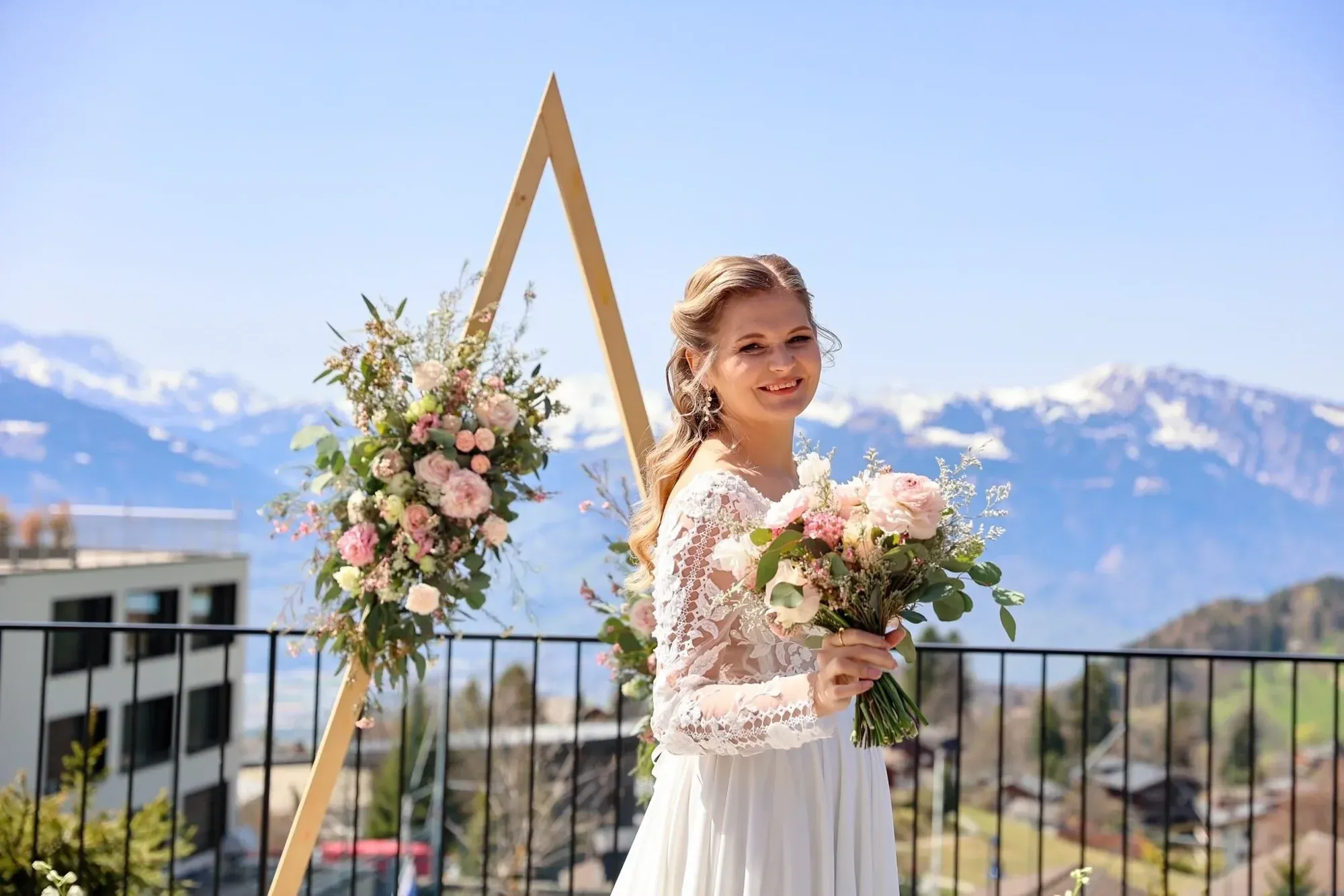 Une femme vêtue d'une robe en dentelle blanche, tenant un bouquet de fleurs roses et blanches, debout en plein air avec des montagnes et un ciel bleu limpide en arrière-plan, et une arche fleurie derrière elle.