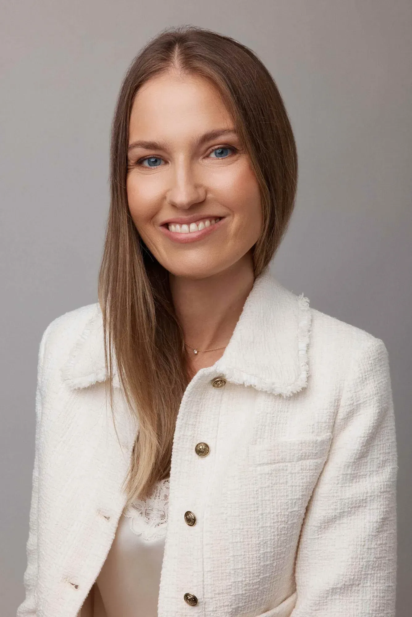 Portrait d'une femme aux longs cheveux châtain clair, aux yeux bleus et à la peau claire, souriante, portant un blazer blanc texturé avec des boutons dorés et un collier délicat, sur un fond gris neutre.