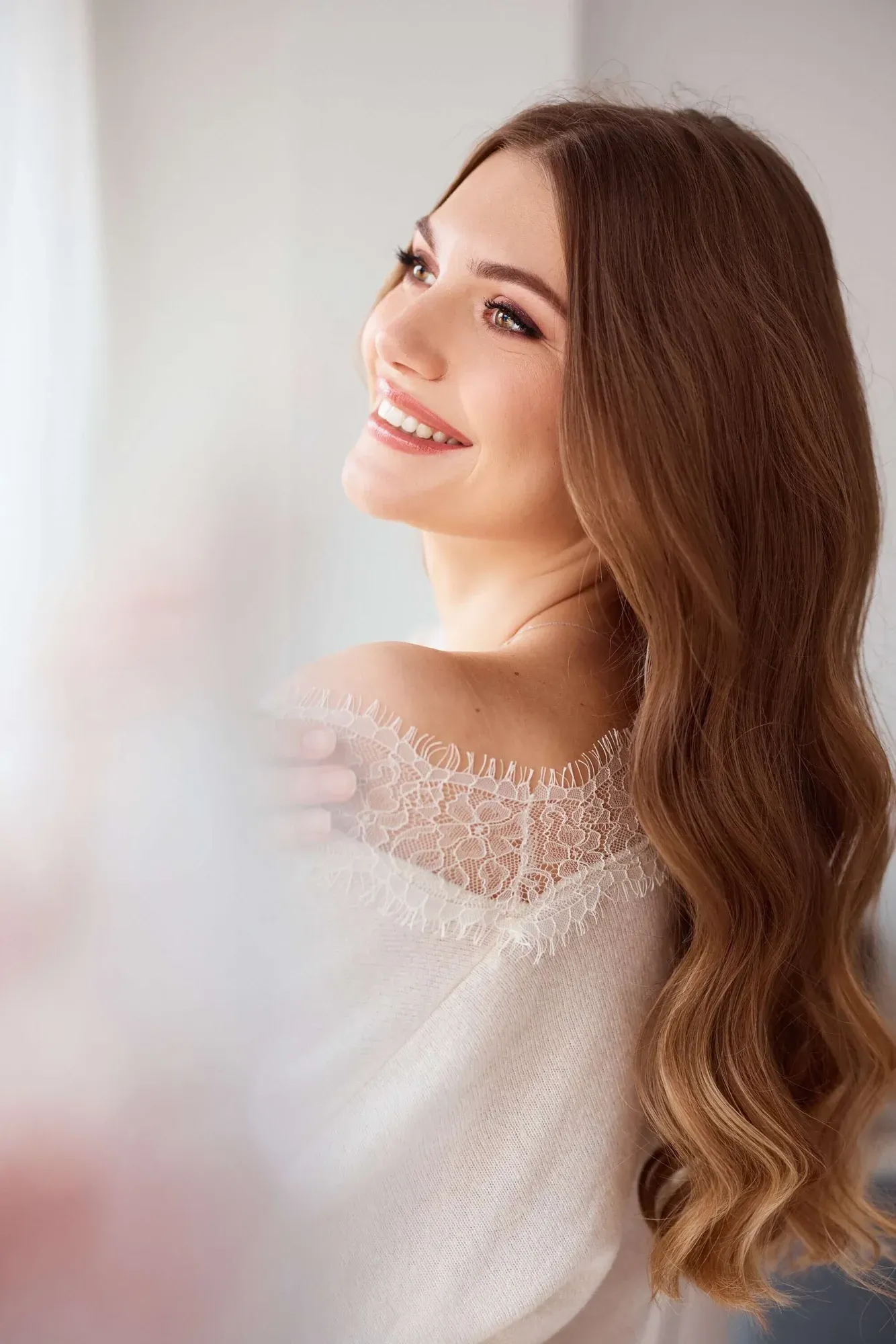 Une femme souriante, aux longs cheveux bruns ondulés et au maquillage discret, vêtue d'un haut en dentelle blanche, debout devant un fond neutre.