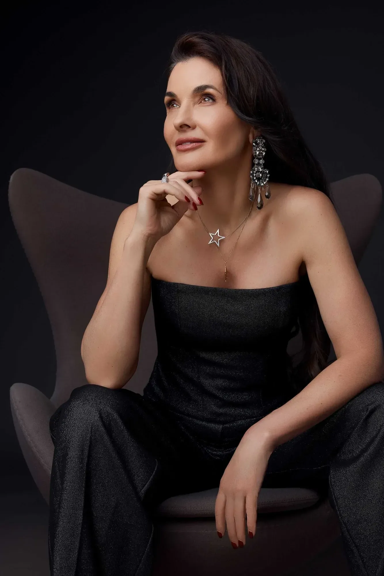 Une femme aux cheveux foncés coiffés en boucles souples, portant de grandes boucles d'oreilles en forme de lustre, plusieurs colliers et pendentifs en forme d'étoile, ainsi qu'une robe noire sans bretelles, assise sur une chaise grise devant un fond noir, le regard tourné vers le haut, l'air pensif.