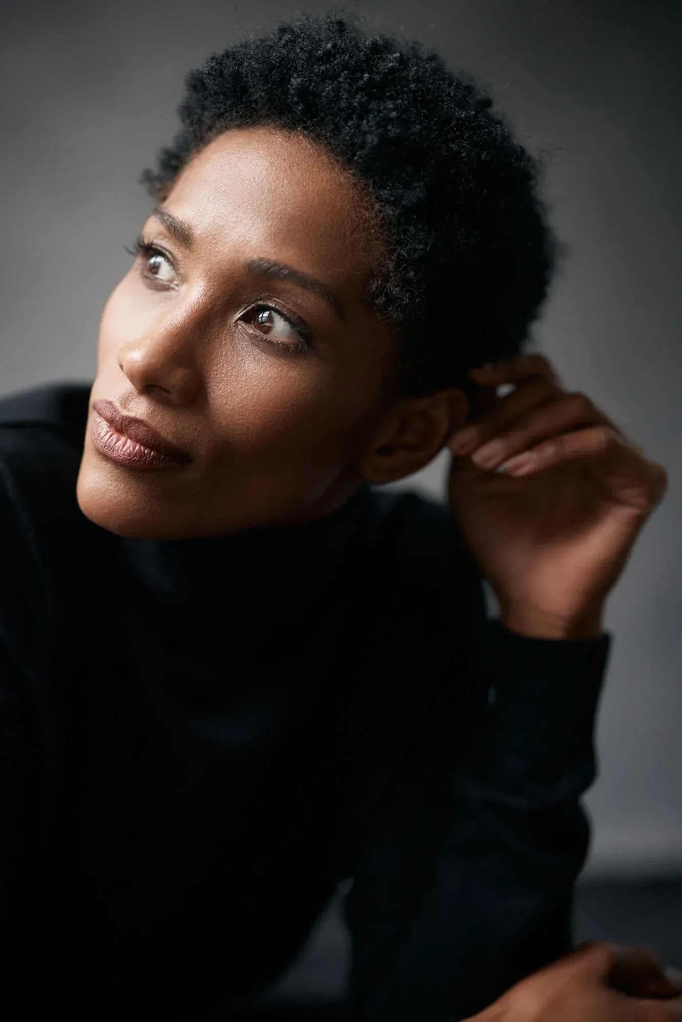 Une femme aux cheveux noirs courts et bouclés regarde légèrement vers le haut et vers la gauche. Elle a la peau mate et porte un haut noir. Sa main droite effleure son oreille et sa tête, et l'arrière-plan est uni et sombre.