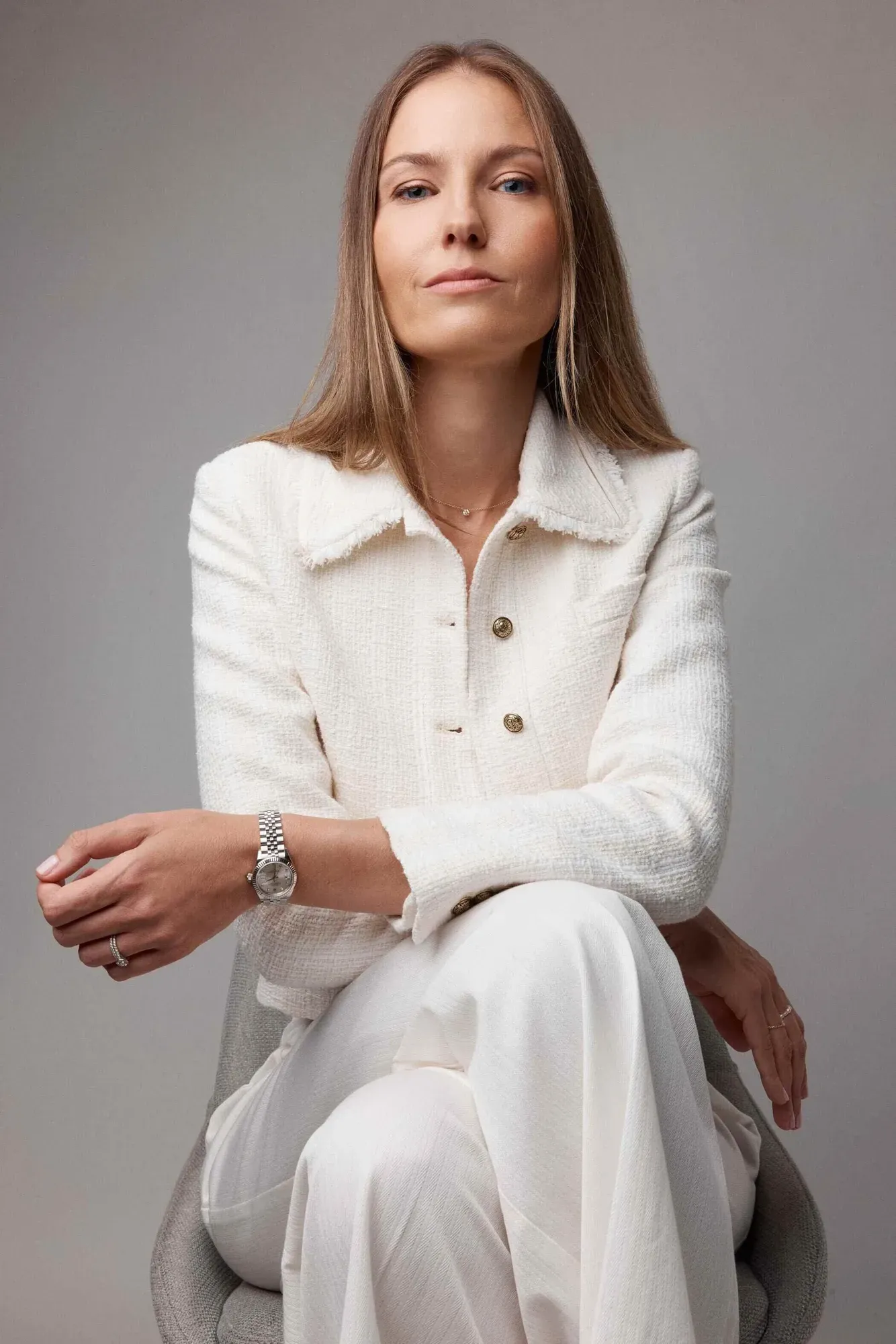 Une femme aux cheveux châtain clair et à la peau claire, portant une veste blanche texturée avec des boutons dorés, est assise sur une chaise grise sur un fond gris uni, regardant l'appareil photo avec assurance.