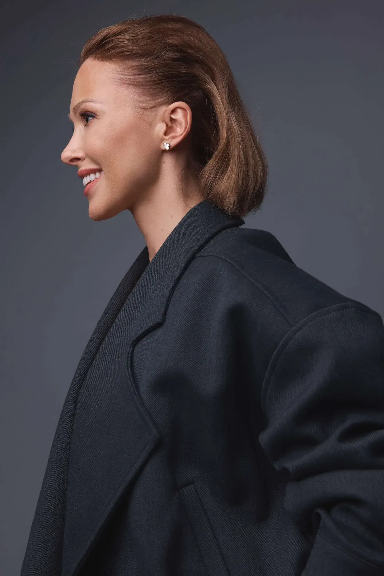 Profil latéral d'une femme aux cheveux courts, châtain clair, souriante, portant un blazer noir et des boucles d'oreilles en perles sur un fond gris foncé.