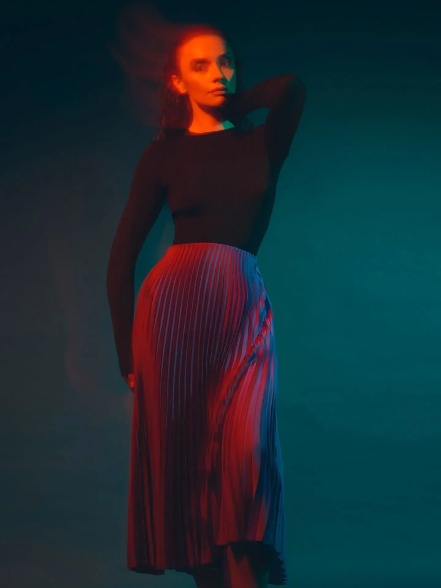 Une femme aux longs cheveux noirs, portant un haut noir et une jupe plissée colorée, debout sur un fond sombre avec des effets de lumière rouge et sarcelle.