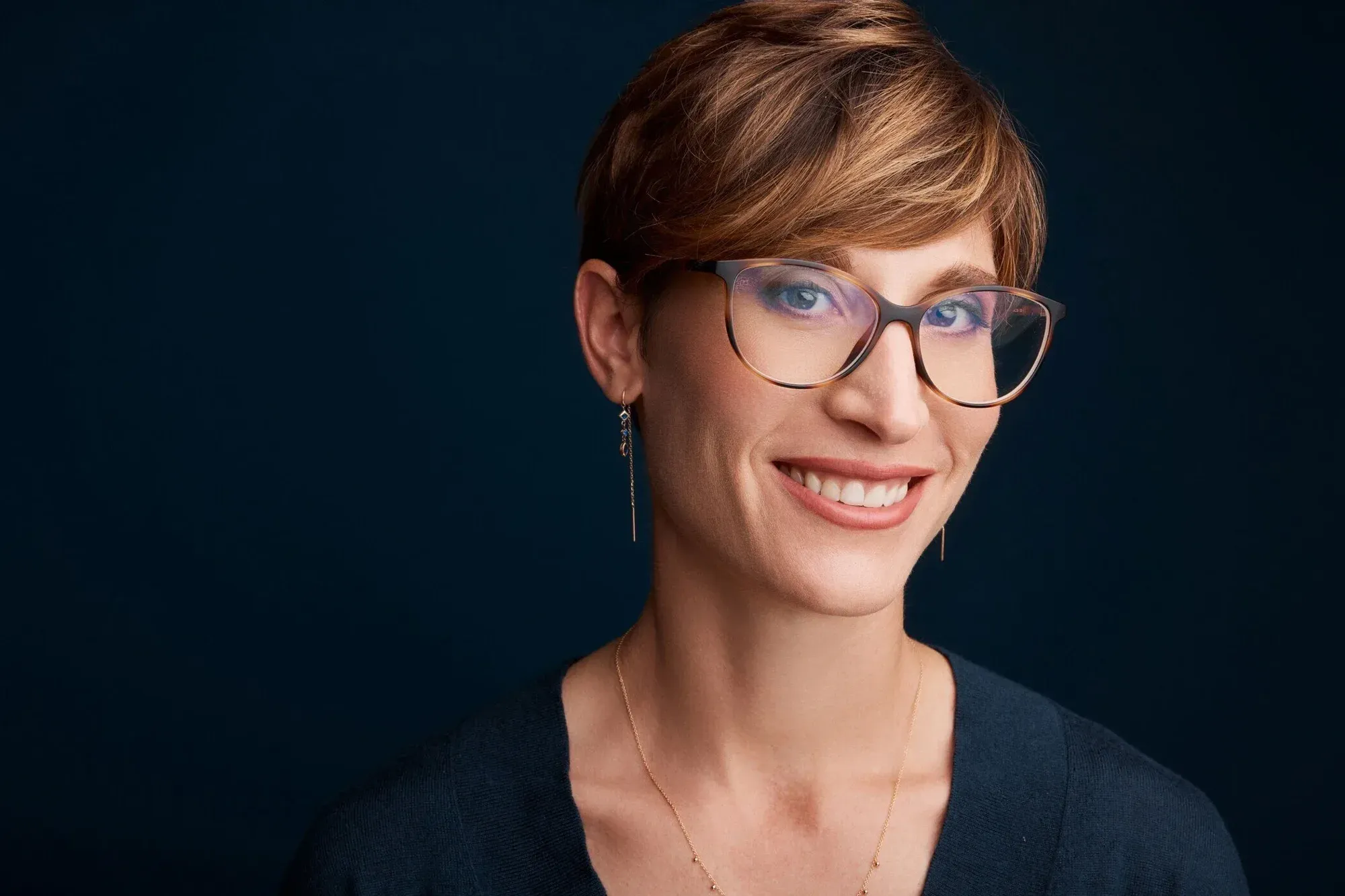 Portrait d'une femme aux cheveux courts brun-rouge, portant des lunettes, des boucles d'oreilles en or et un collier en or, souriant sur un fond bleu foncé.