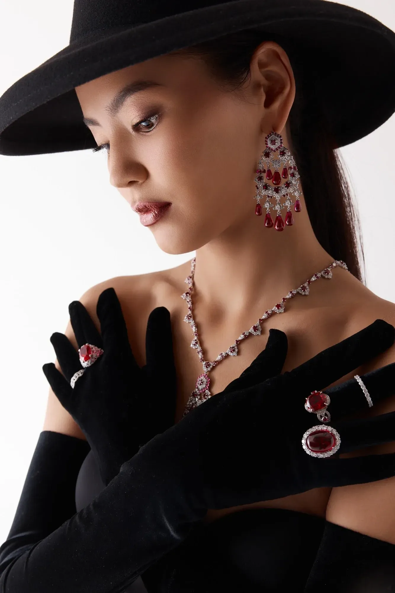 Une femme portant un chapeau noir à large bord, des gants noirs et une robe noire, agrémentée de bijoux rouges et argentés, notamment un collier, des boucles d'oreilles, des bagues, des bracelets et une broche. Elle regarde vers le bas, l'air impassible.