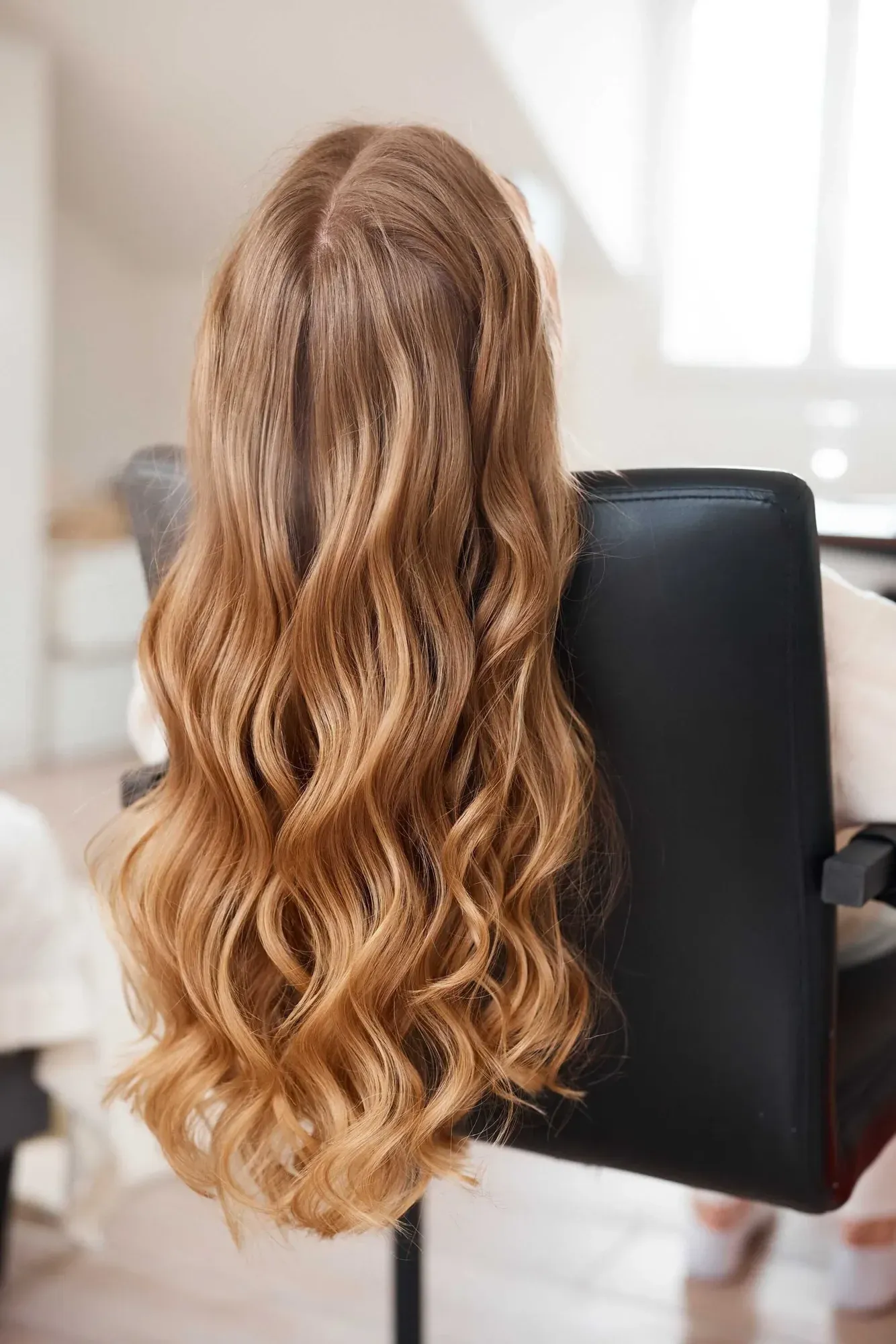 De longs cheveux blonds ondulés appartenant à une personne assise sur une chaise noire dans une pièce lumineuse aux murs blancs et aux grandes fenêtres.