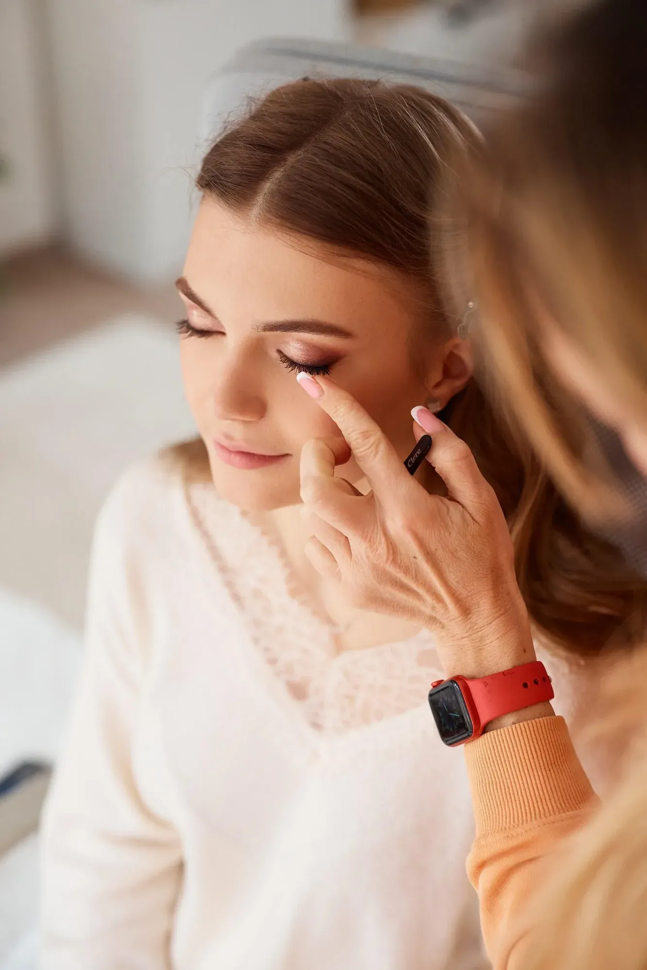Une femme se maquille, les yeux fermés, en effleurant délicatement sa paupière du doigt. Elle a de longs cheveux châtains, le teint clair, et porte un haut en dentelle blanche. Une personne portant une montre connectée rouge et une manche orange l'aide en lui tenant la paupière.