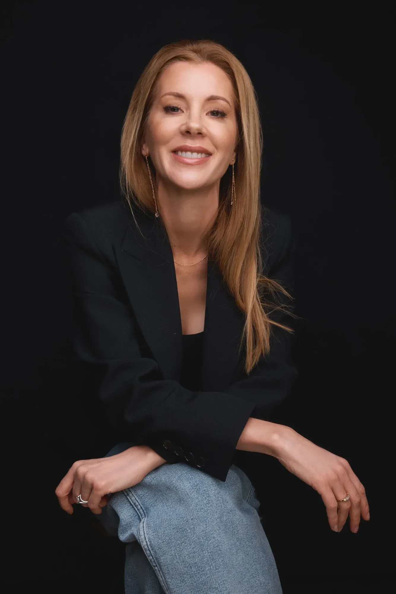 Une femme aux longs cheveux roux ondulés sourit devant un fond noir. Elle porte un blazer noir, un jean bleu clair et des bijoux en or.
