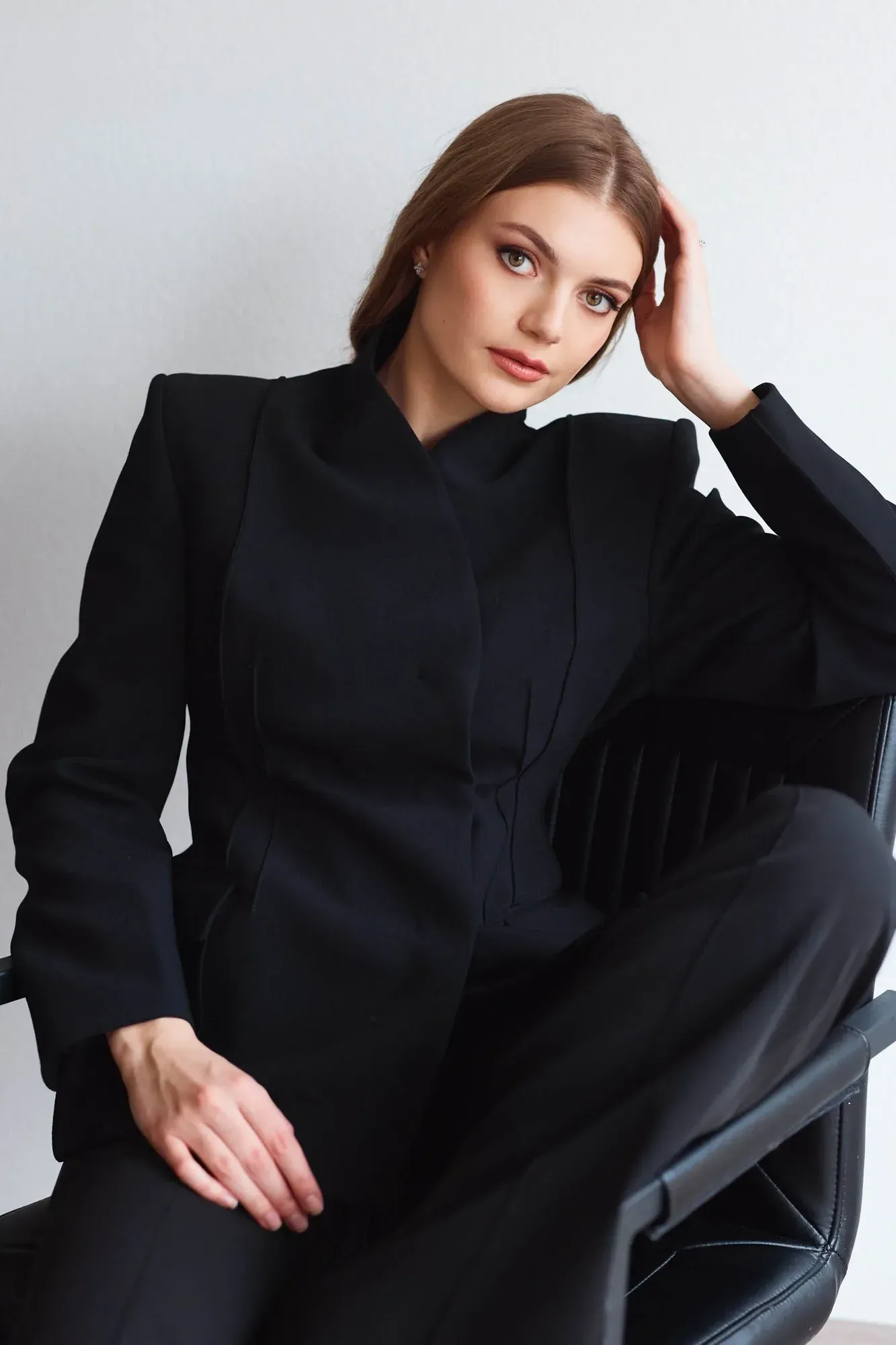 Une femme aux cheveux châtain clair mi-longs, vêtue d'un blazer noir, assise sur un fauteuil en cuir noir, se caressant les cheveux, l'air impassible.