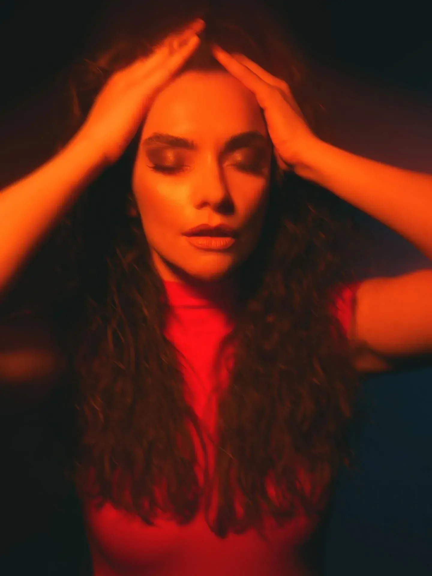 Une femme aux cheveux bouclés et maquillée, les yeux fermés, les mains sur la tête.