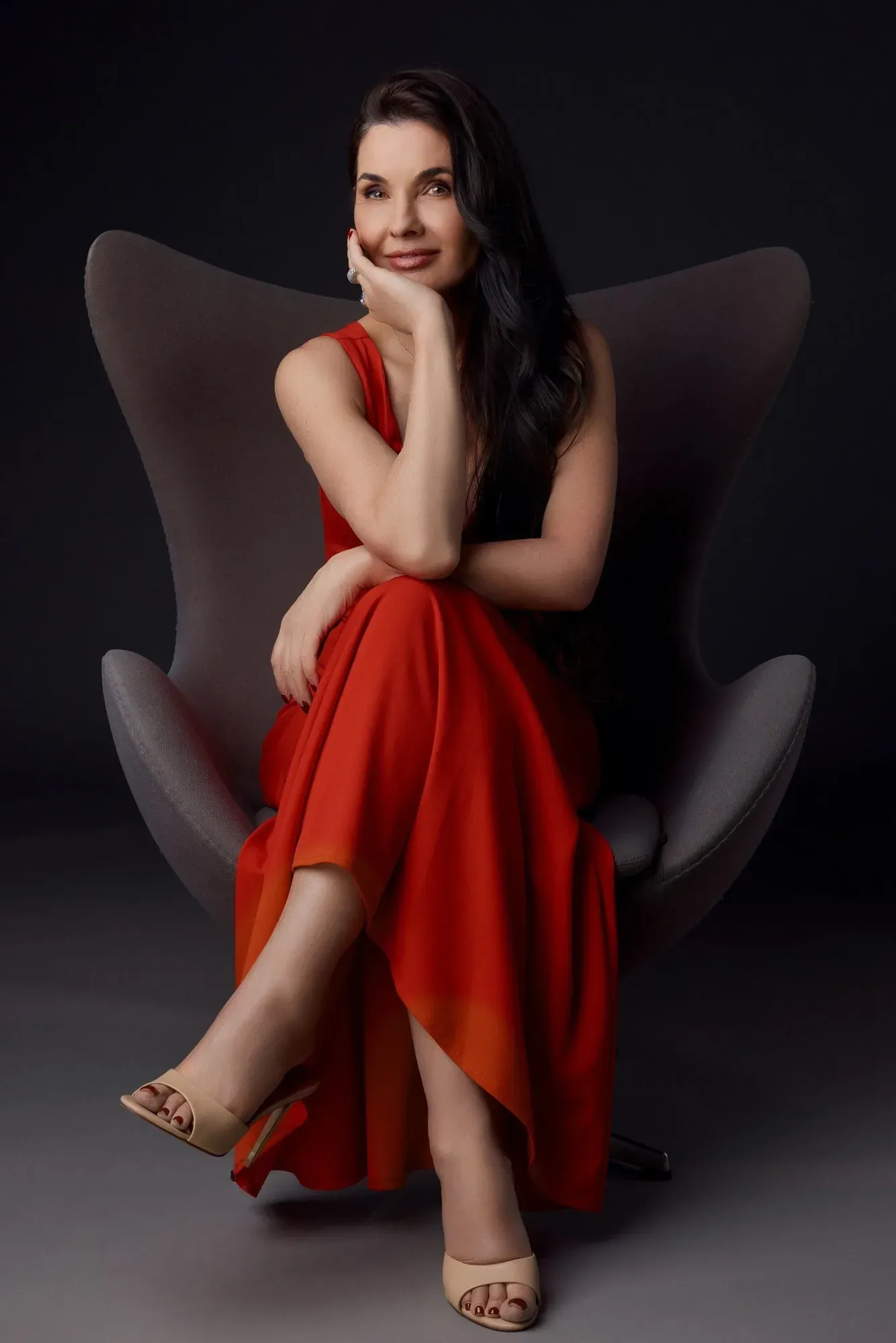 Une femme en robe rouge, assise dans un fauteuil moderne gris à haut dossier, posant, une main sur le visage, l'air sûr d'elle, sur fond noir.