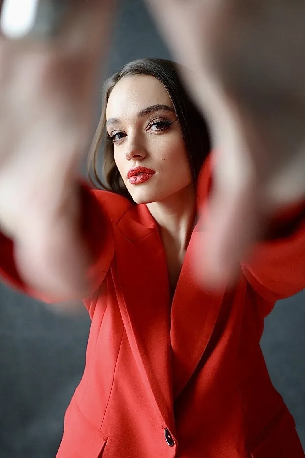 Une femme aux cheveux bruns, au rouge à lèvres rouge et au blazer rouge, regardant la caméra de face, les mains levées vers la caméra.