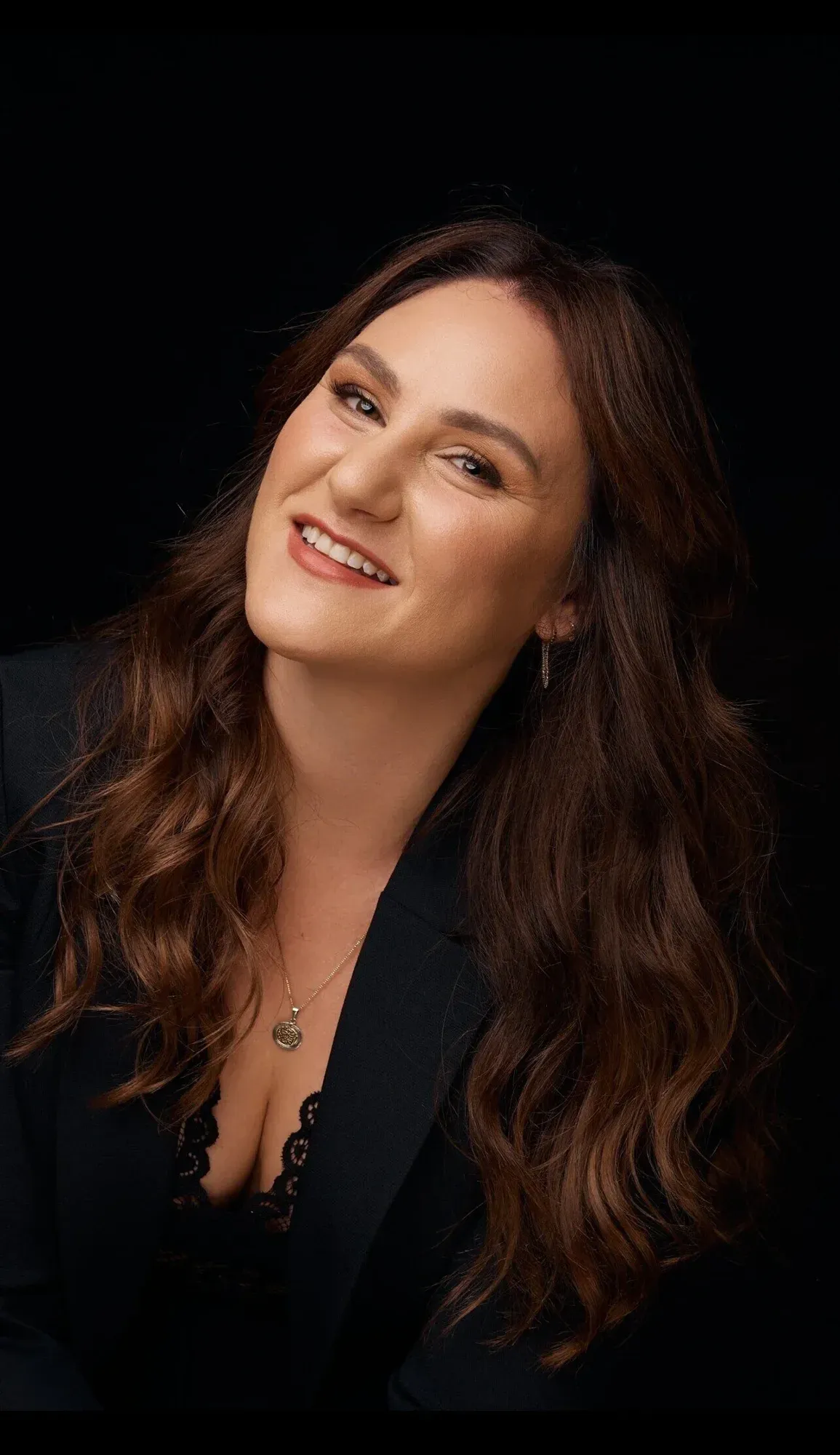 Une femme aux longs cheveux bruns ondulés souriant et faisant un clin d'œil à l'appareil photo, portant un blazer noir et un collier sur un fond noir.