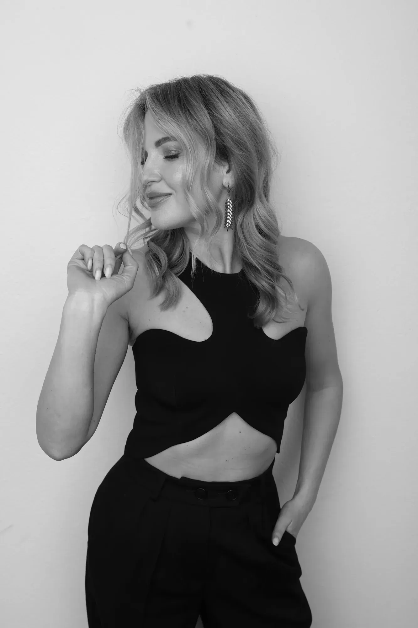 Une femme aux cheveux blonds ondulés, vêtue d'un haut noir sans manches avec une découpe et d'un pantalon noir taille haute, debout contre un mur uni. Elle porte des boucles d'oreilles et sourit les yeux fermés, tenant une mèche de cheveux d'une main.